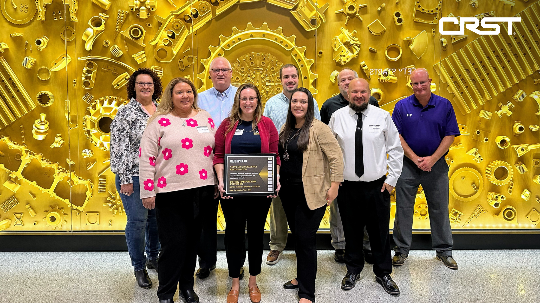 A group of CRST team members stand in front of a gold, gear-themed wall, proudly holding a Supplier Excellence Recognition award from Caterpillar. The award highlights CRST’s outstanding service and commitment as a top-tier supplier.