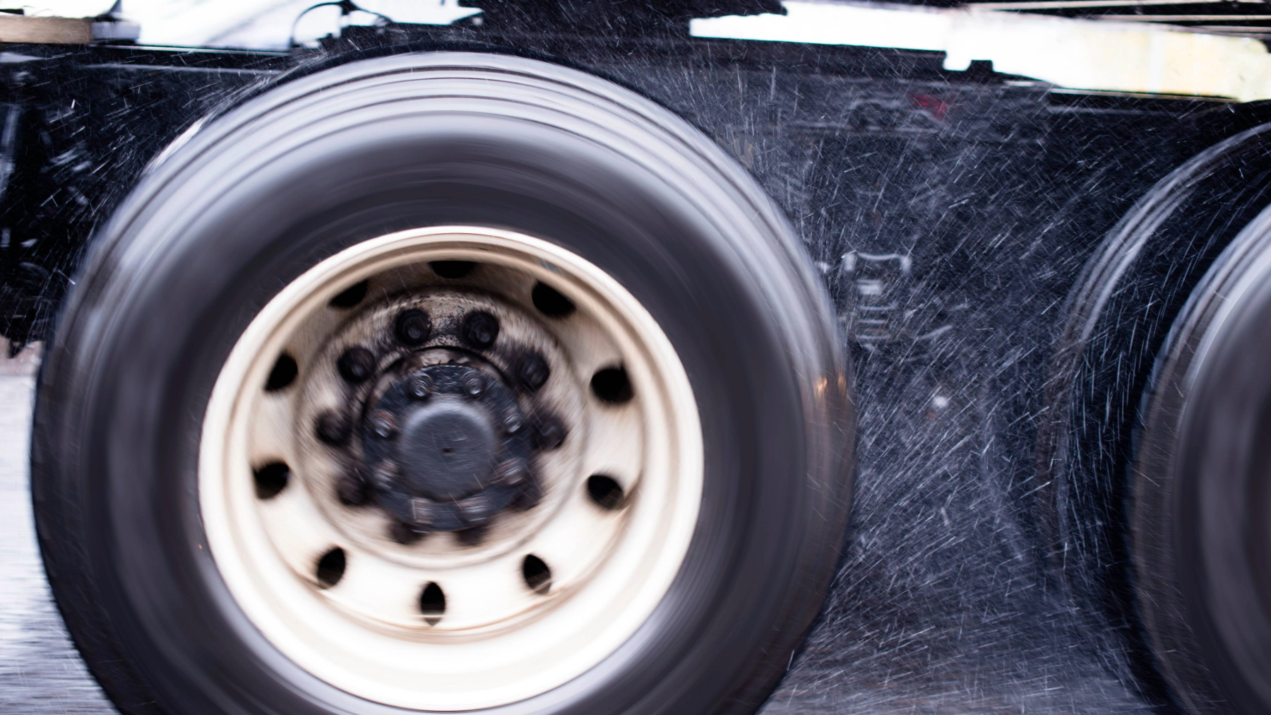 A close-up of a spinning semi-truck tire on a wet road, with water spraying from the motion. The blurred effect emphasizes speed and movement, highlighting the risks of hydroplaning in wet conditions.