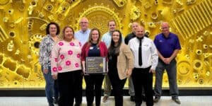 A group of CRST team members stands together in front of a striking gold industrial-themed backdrop, celebrating their Caterpillar Supplier Excellence Certification. Kara Hinze, director of operations, holds the award plaque, recognizing the team’s outstanding performance and leadership in logistics.