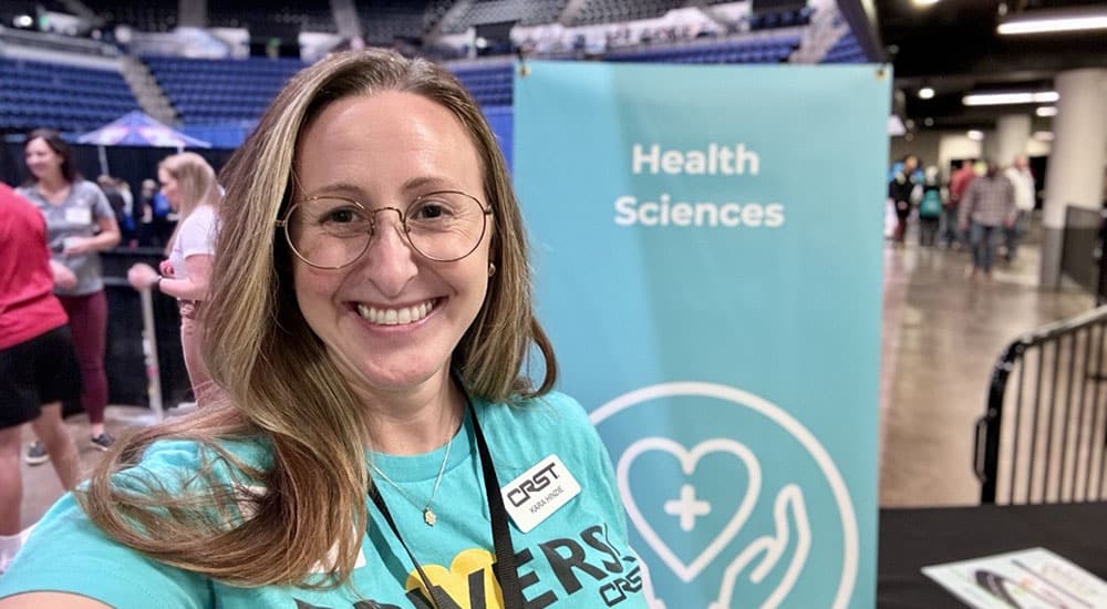 CRST’s Kara Hinze smiles at a career fair, wearing a "DRIVERS" t-shirt and name badge. Behind her, a "Health Sciences" banner is visible, indicating the event’s focus. This image accompanies the blog "Redefining Logistics Leadership: CRST’s Kara Hinze Transforms a Dedicated Fleet.