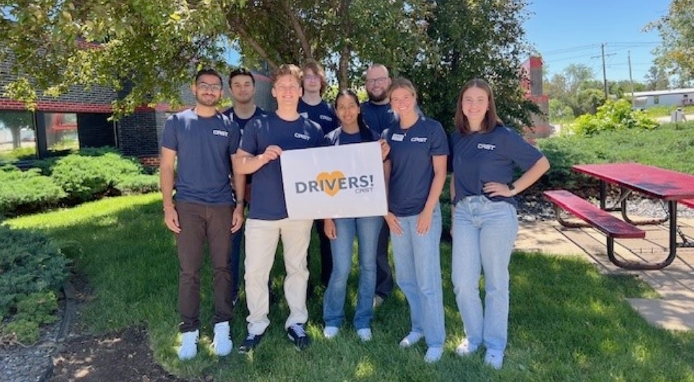 A group of CRST interns and early career team members stand together outdoors, smiling and holding a sign that reads "DRIVERS! CRST." The team is celebrating CRST’s recognition as a 2025 Handshake Early Talent Award winner.